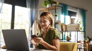 Frau sitzt zu Hause am Tisch mit offenem Laptop und Smartphone in der Hand